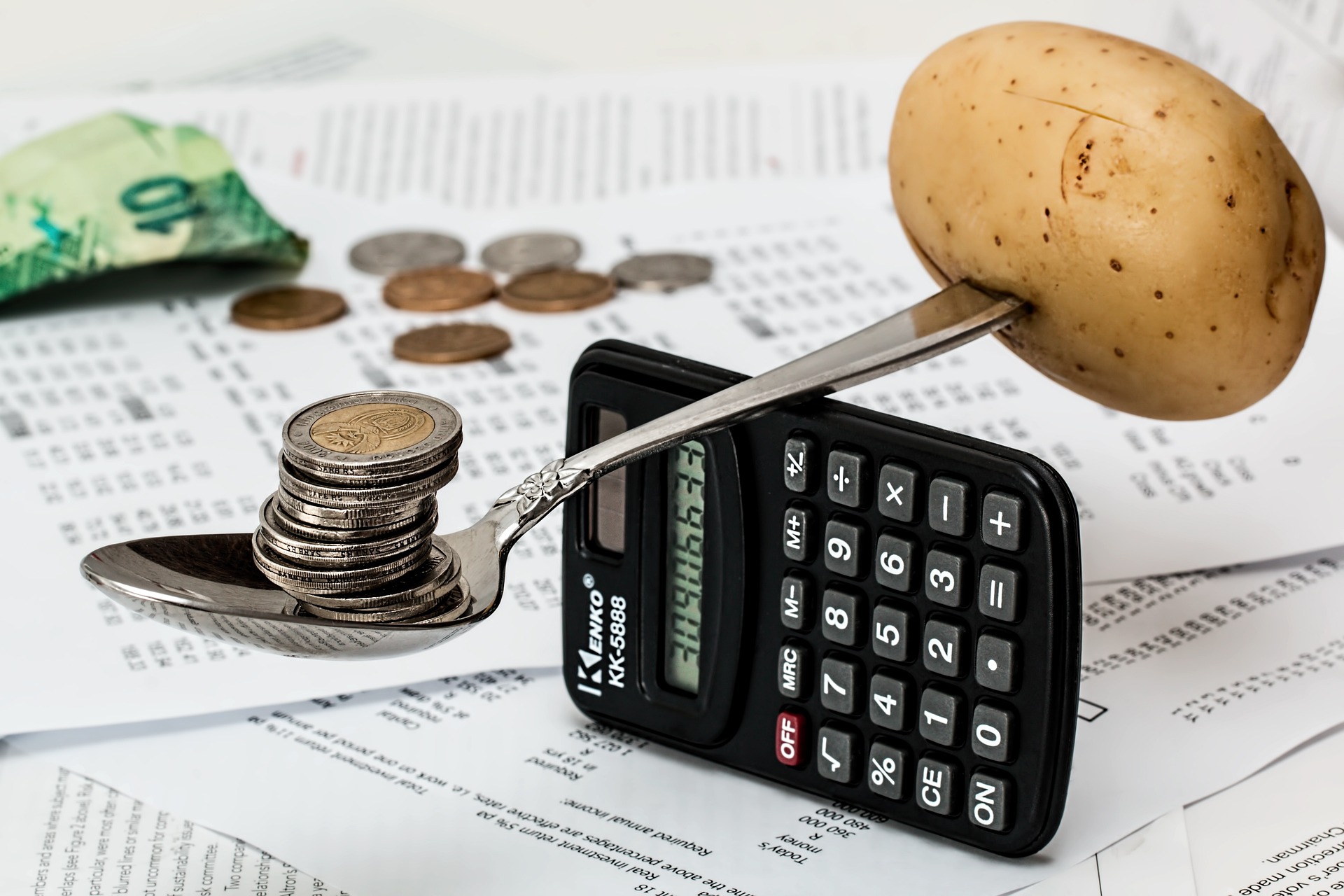 Pieces of white A4 paper with columns of numbers all over them blurred into the background with some cash and coins on them. In the foreground is a calculator balancing on its side with a spoon sitting across the top with some coins balancing equally on one side against a potato on the other side.